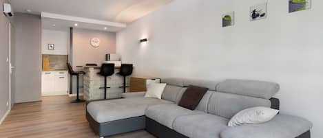 Una sala moderna con un sofá seccional gris, una cocina al fondo y una mesa de comedor con sillas.