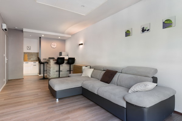 A modern living room with a grey sectional sofa, a kitchen area in the background, and a dining table with chairs. - Refuge des tribus Apartment: Mountain View, Shared Garden, Private Balcony (Villard-de-Lans)