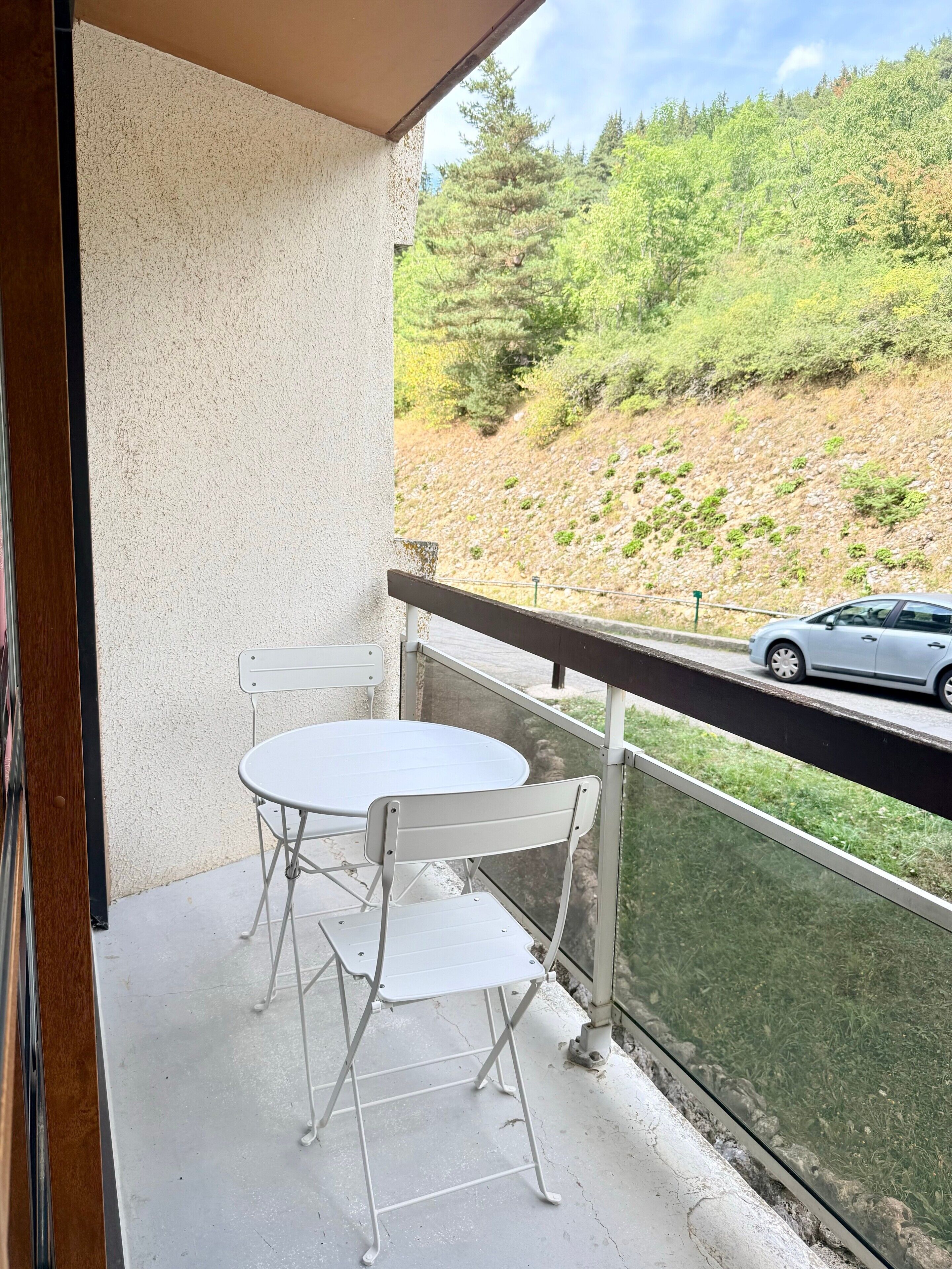 Un balcón con una mesa y sillas blancas, con vistas a una carretera y una colina con árboles.