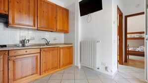 A kitchen with wooden cabinets, a built-in oven, a sink, and a wall-mounted television. - Three-Room Apartment with Garden 100 Meters from the Beach - Sole e Mare Tourist Village (Rio)