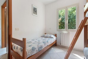 A single bed with a floral bedspread in a room with white walls, a window with a view of greenery, and a radiator. - Three-Room Apartment with Garden 100 Meters from the Beach - Sole e Mare Tourist Village (Rio)