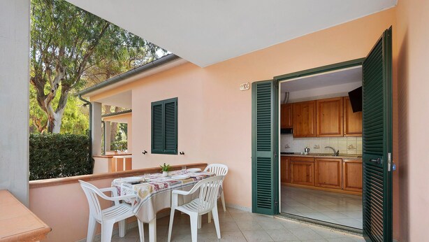 An outdoor dining area with a table and chairs, a view of the kitchen through an open door, and a tree in the background. - Three-Room Apartment with Garden 100 Meters from the Beach - Sole e Mare Tourist Village (Rio)