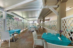 An outdoor patio area with tables and chairs, covered by a white roof, and surrounded by white lattice fencing. - Three-Room Apartment with Garden 100 Meters from the Beach - Sole e Mare Tourist Village (Rio)