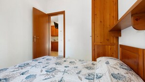 A bedroom with a bed, wooden wardrobe, and a kitchen visible through an open door. - Three-Room Apartment with Garden 100 Meters from the Beach - Sole e Mare Tourist Village (Rio)