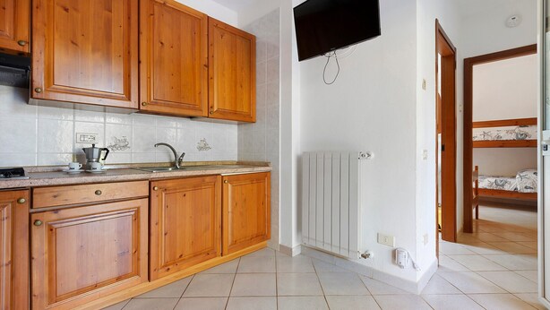 A kitchen with wooden cabinets, a built-in oven, a sink, and a wall-mounted television. - Three-Room Apartment with Garden 100 Meters from the Beach - Sole e Mare Tourist Village (Rio)