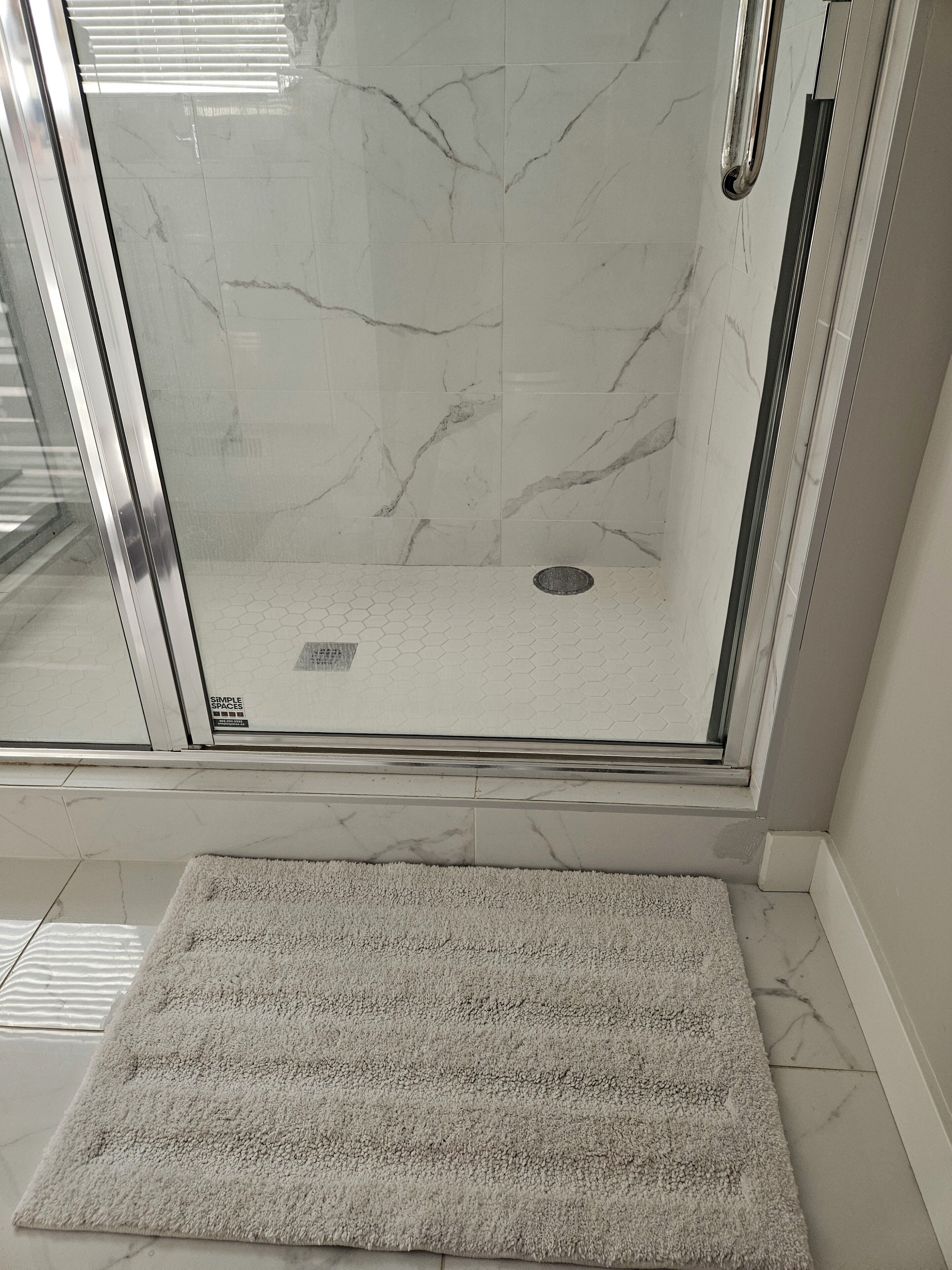 A glass-enclosed shower with a marble floor and a textured rug in front.
