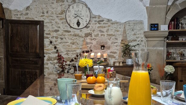 A rustic dining area with stone walls, a wooden table set with breakfast items, and a clock on the wall. - Épinette Guest Room with Shared Terrace, Garden, and Non-Private Jacuzzi (Vellerot-lès-Vercel)