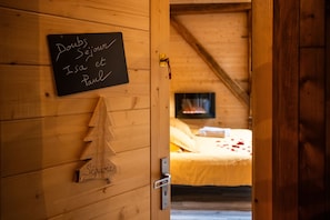 Un interior de madera con una pizarra mostrando nombres, un pequeño abeto navideño y una cama con una colcha amarilla y pétalos de rosa.