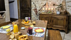 A rustic dining area with a wooden table set for breakfast, including bread, fruit, and a beverage. A clock is mounted on the wall, and a potted plant is on a wooden cabinet.