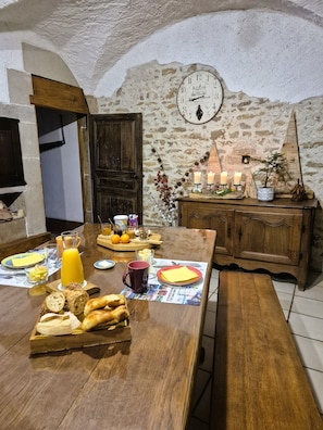 A rustic dining area with a wooden table set for breakfast, including bread, fruit, and a beverage. A clock is mounted on the wall, and a potted plant is on a wooden cabinet.