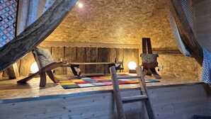 A wooden loft room with a sloped ceiling, a colorful rug, a ladder, and a chair.
