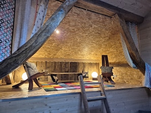 A wooden loft room with a sloped ceiling, a colorful rug, a ladder, and a chair.