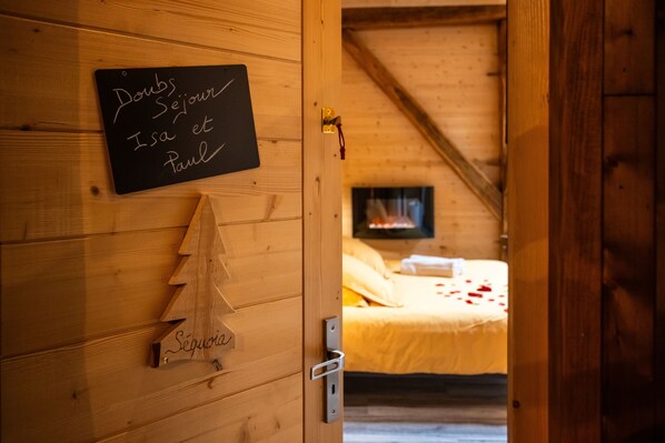 A wooden interior with a chalkboard displaying names, a small wooden Christmas tree, and a bed with a yellow blanket and rose petals.