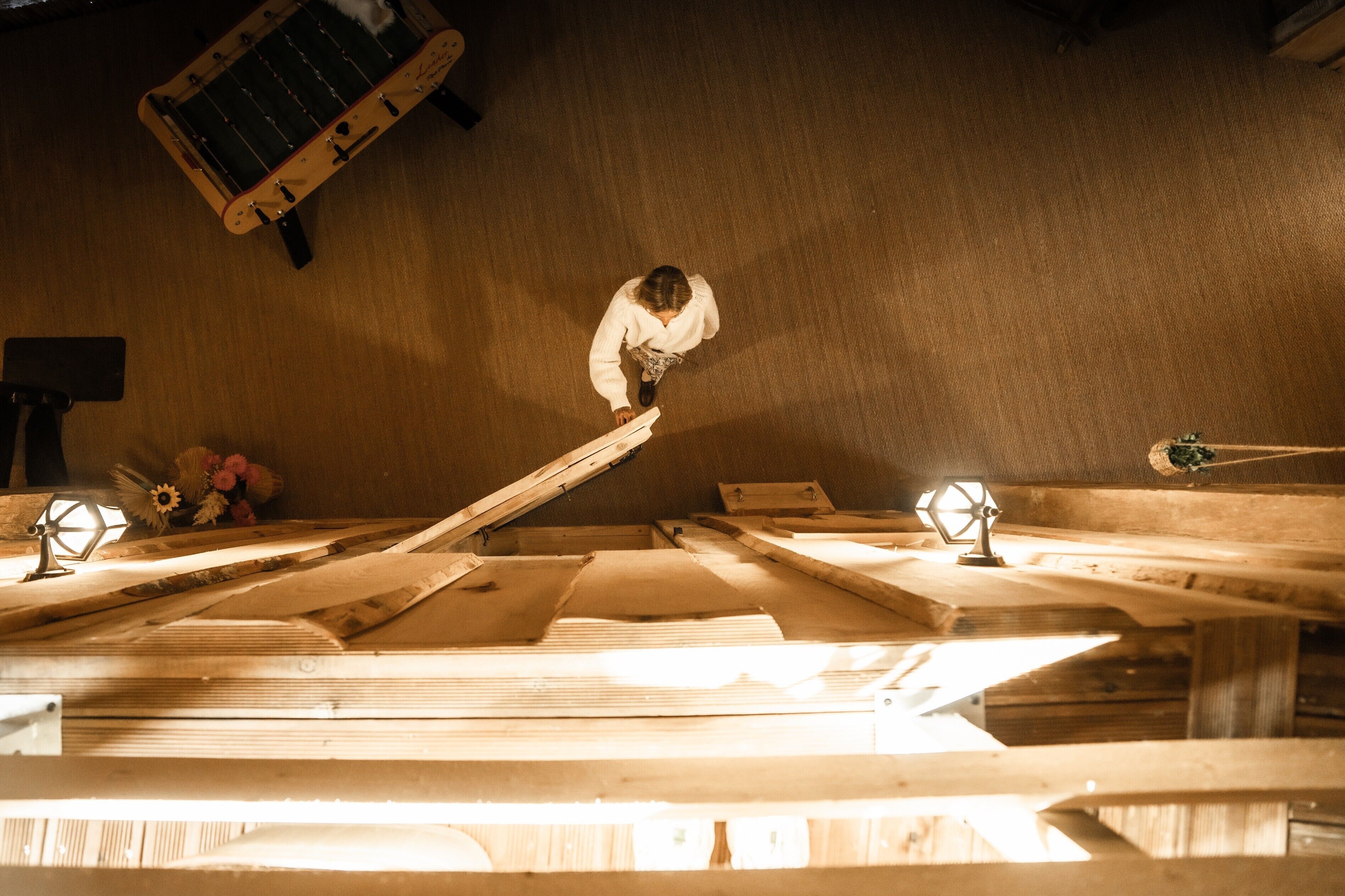 Een werkplaats met houten banken en gereedschap, een persoon werkt aan een bank, en een hanglamp.