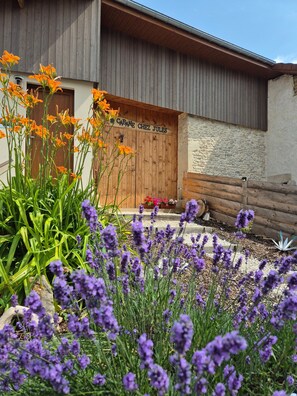 Una puerta de madera con una placa, un muro de piedra y un jardín con lavanda y flores de naranja.