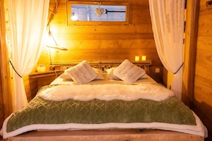 A wooden room with a bed, curtains, a window, and a nightstand with candles.
