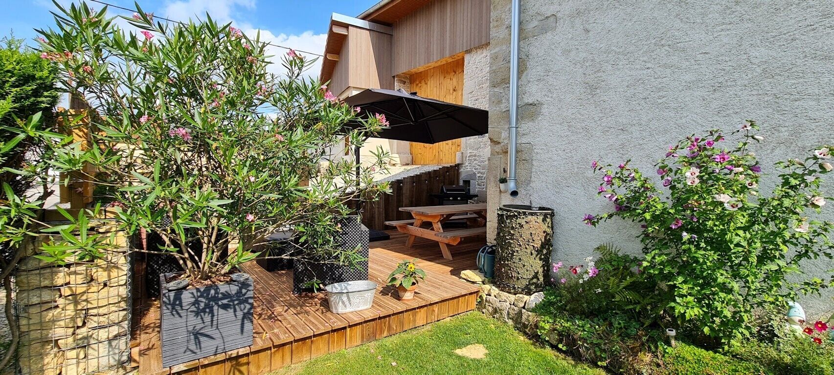Un patio trasero con terraza de madera, zona de barbacoa y plantas en macetas.