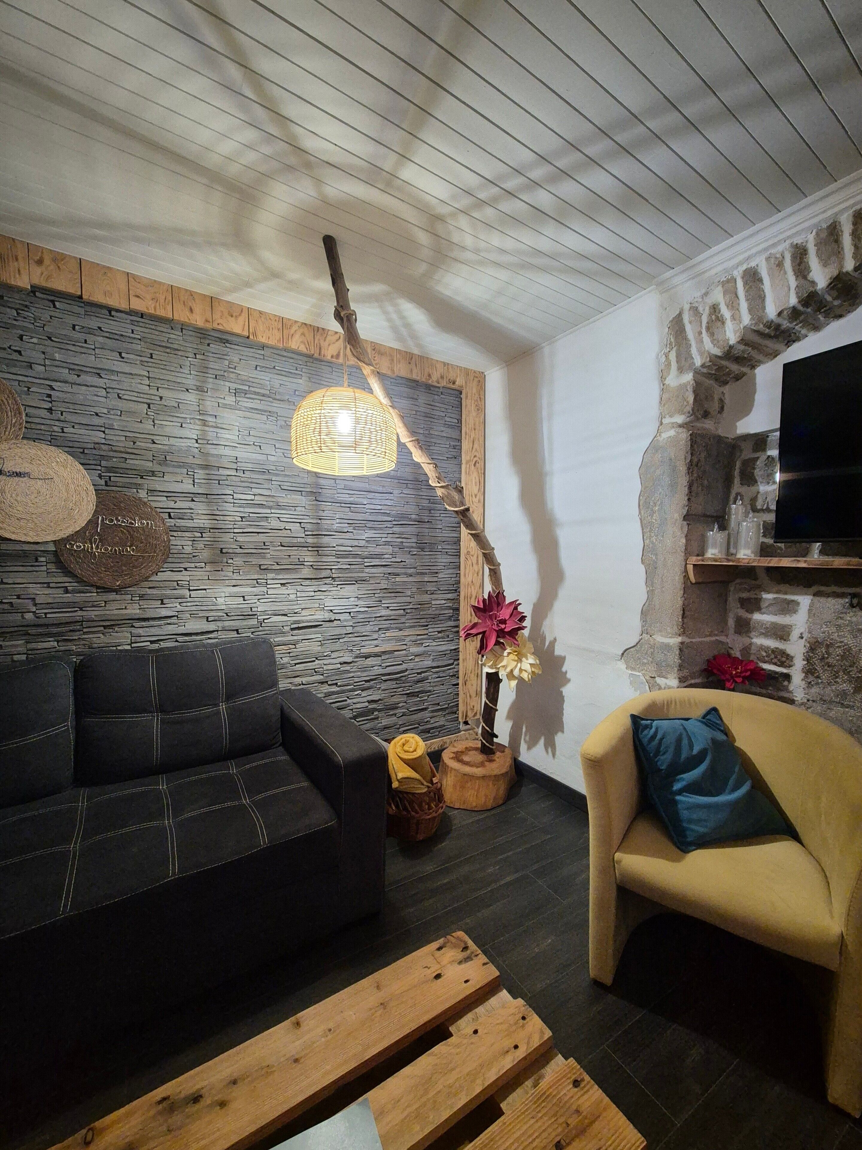 Een woonkamer met een stenen muur, een houten plafond, een zwarte bank, een gele fauteuil, een houten salontafel en een hanglamp.
