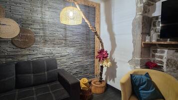 A living room with a stone wall, a wooden ceiling, a black sofa, a yellow armchair, a wooden coffee table, and a hanging light fixture.