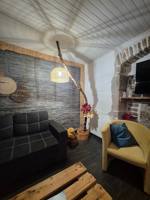 A living room with a stone wall, a wooden ceiling, a black sofa, a yellow armchair, a wooden coffee table, and a hanging light fixture. - Gîte Chez Jules with Spa, Private Terrace, and Wi-Fi (Vellerot-lès-Vercel)