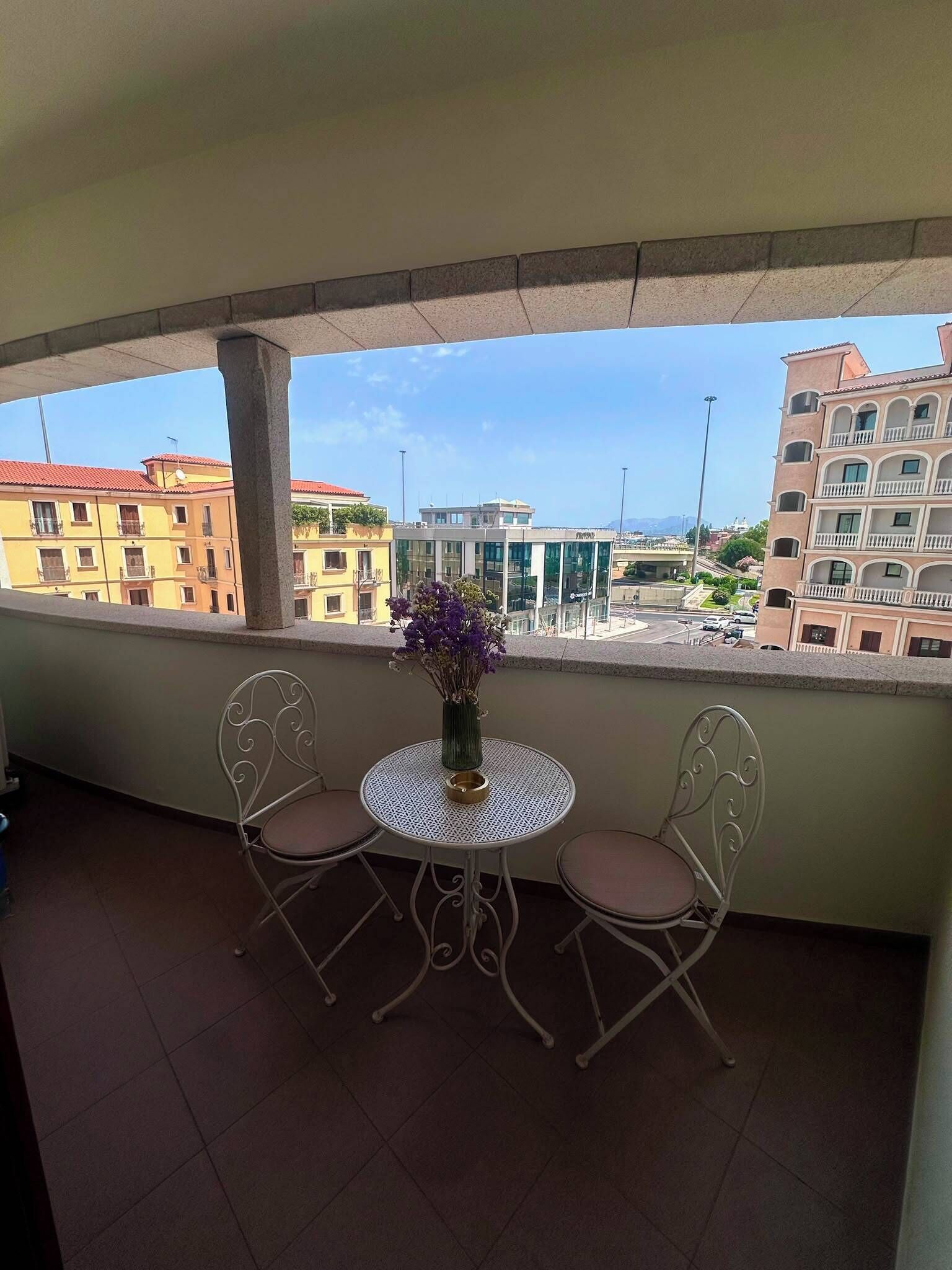 Un balcone con due sedie e un tavolo, affaccia su un panorama urbano con edifici e cielo sereno.