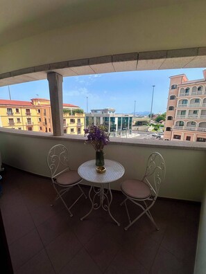 Un balcone con due sedie e un tavolo, affaccia su un panorama urbano con edifici e cielo sereno.