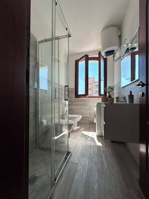 Un bagno moderno con box doccia in vetro, un water, un lavandino e un'ampia finestra con vista sugli edifici.