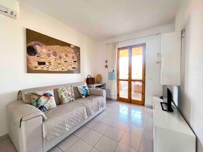 Ein Wohnzimmer mit einem beigefarbenen Sofa, bunten Kissen, einem großen abstrakten Gemälde, einem Fernseher und einer Glastür zum Garten.