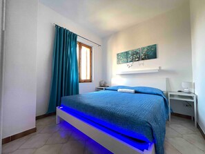 Ein Schlafzimmer mit einem Bett, einem Nachttisch, einer Lampe, einem Fenster mit Vorhängen und einem an der Wand befestigten Kunstwerk.
