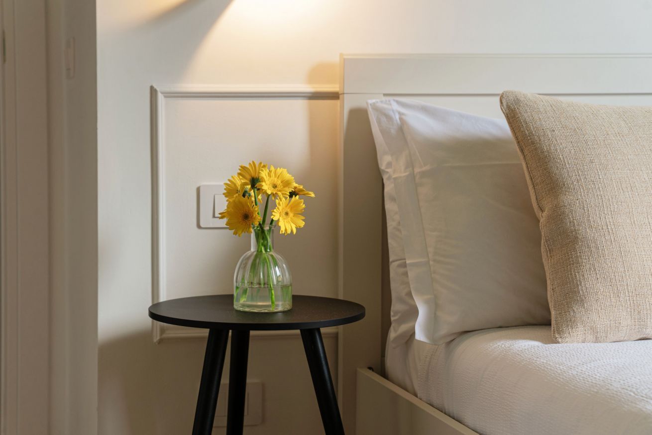 A bed with a white headboard and a beige pillow, a small round table with a clear vase of yellow flowers, and a wall-mounted light fixture.
