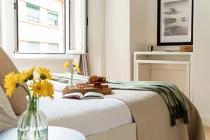 Ein Schlafzimmer mit einem Bett, einem Nachttisch mit Buch und Blumen, einem Fenster mit Blick auf Gebäude und einem gerahmten Bild an der Wand.