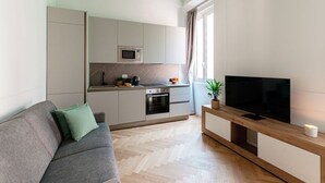 A modern living space with a sofa, a flat-screen TV, a kitchen area, and a window with curtains. - Incanto 107 - My Home in Como (Como)