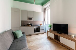 A modern living space with a sofa, a flat-screen TV, a kitchen area, and a window with curtains. - Incanto 107 - My Home in Como (Como)