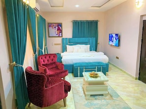 Habitación de hotel con cama, un sillón rojo, una mesita con un cuenco y cortinas azul verdosas.