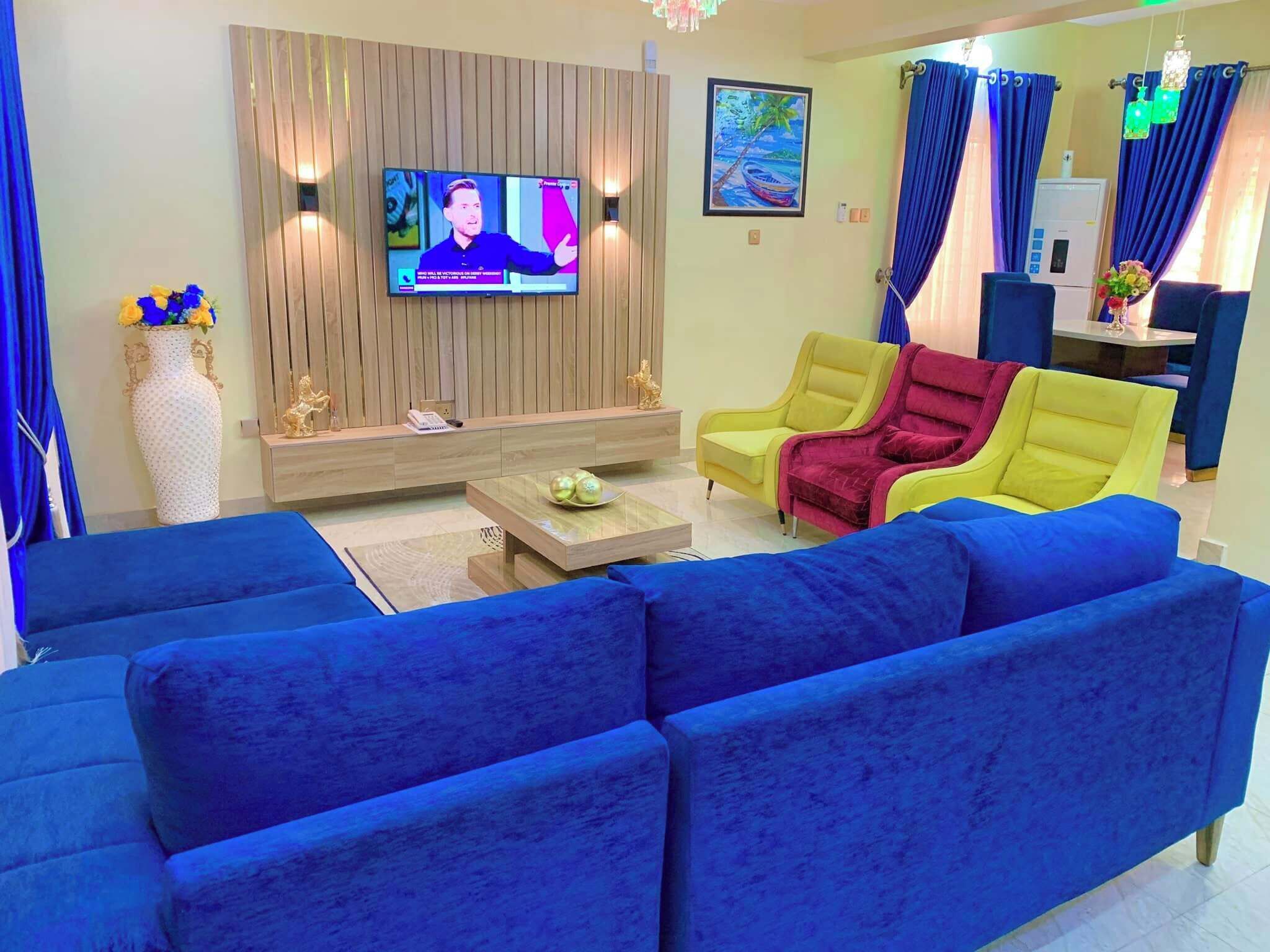 A living room with a large blue sofa, a yellow armchair, a TV mounted on a wooden wall, and a dining area with blue curtains.