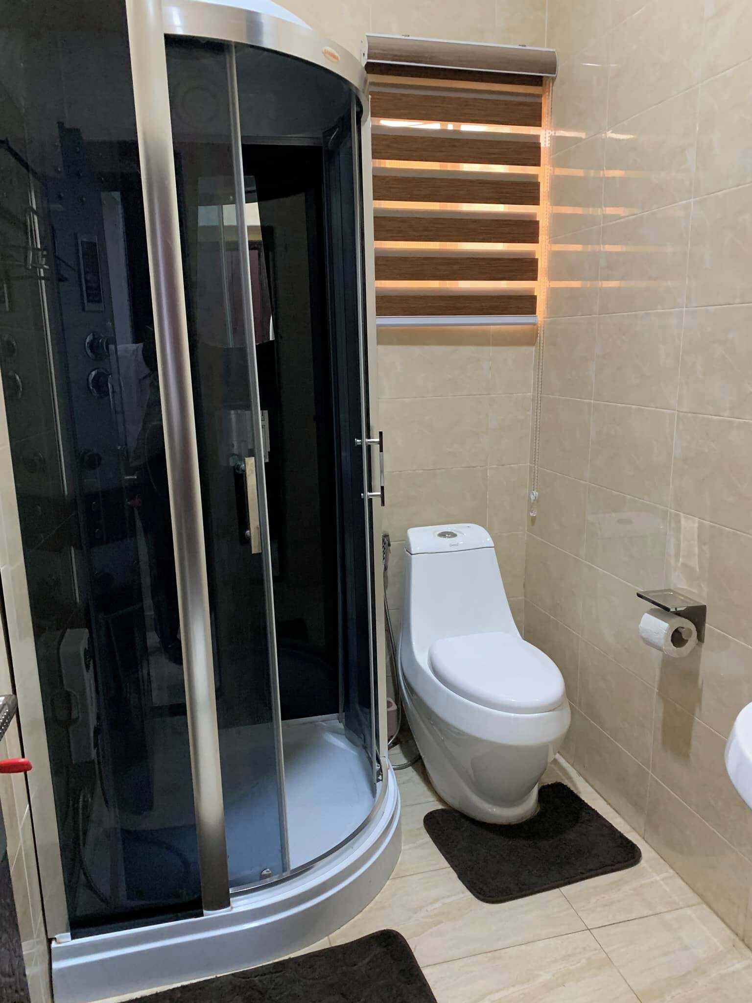 Un baño con ducha envuelta en vidrio, inodoro y ventana con persianas.