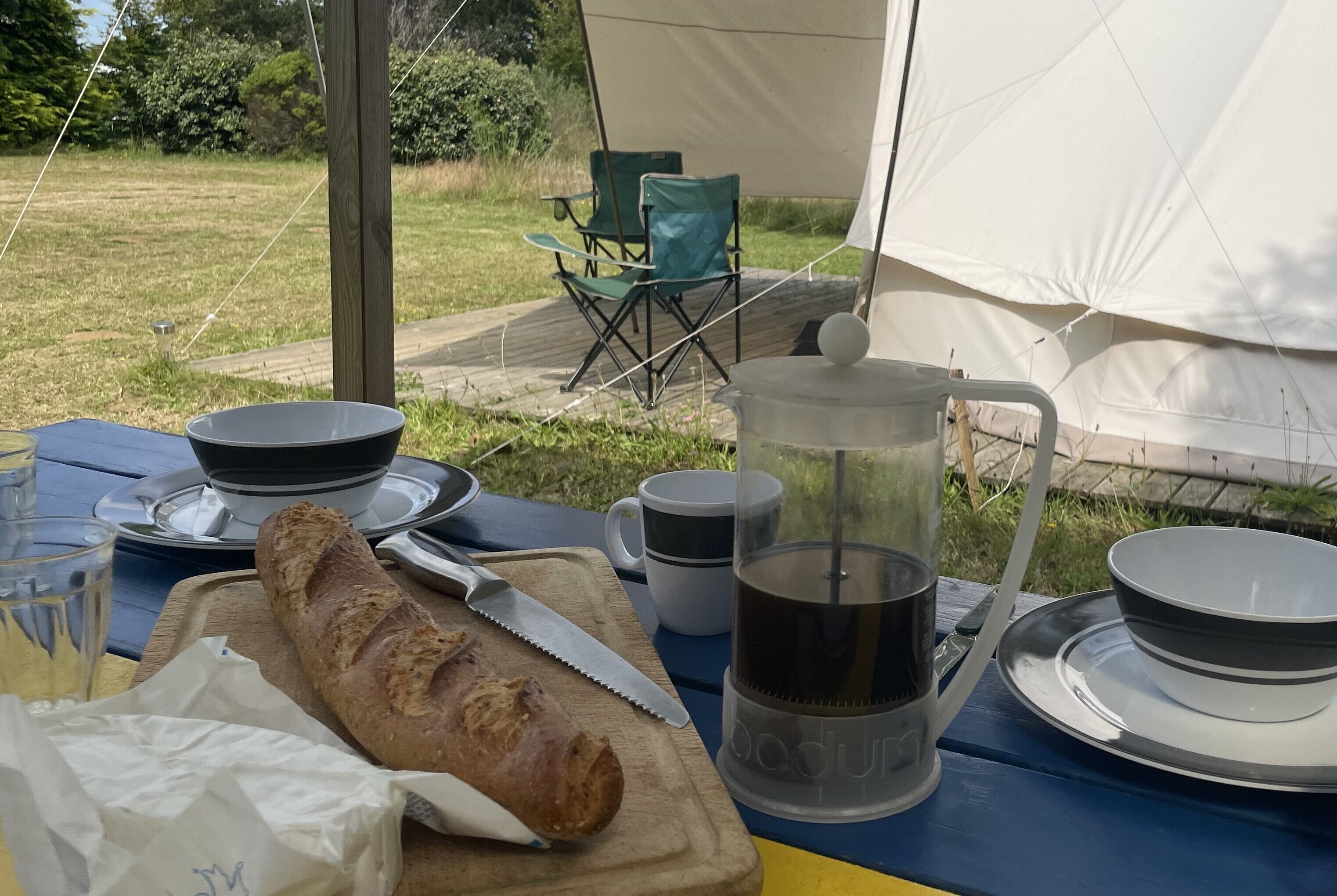 A campsite with a table set for a meal, including a baguette, a coffee pot, and various dishes.