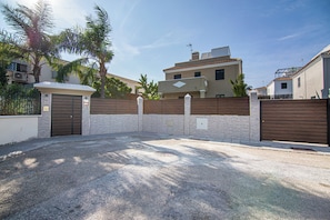 A gated parking area with a gravel surface, a metal gate, and a building with a tiled roof in the background.
