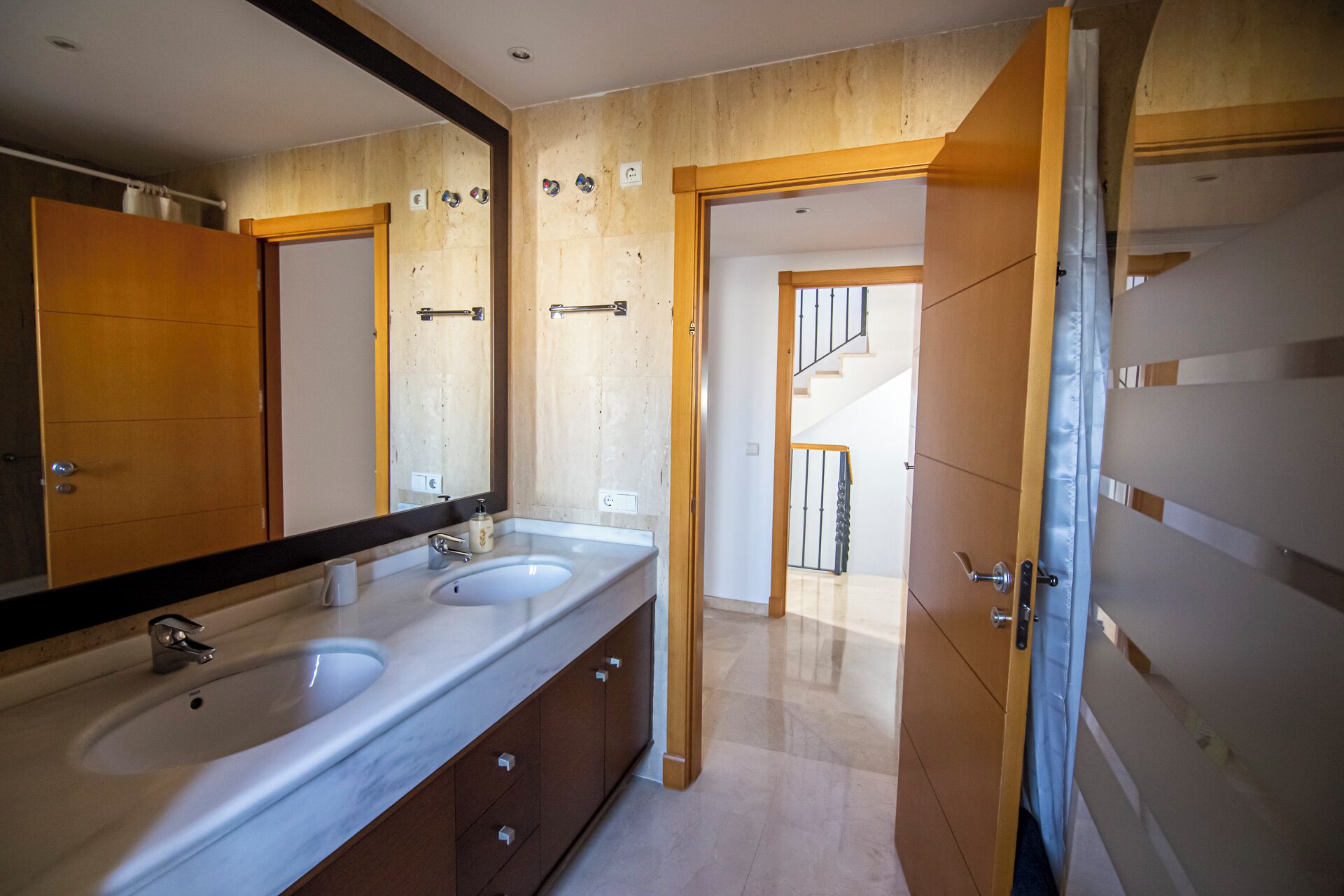A bathroom with a double sink, a large mirror, wooden paneling, and a view of a staircase leading to another room.