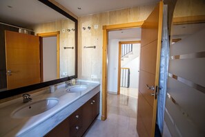 A bathroom with a double sink, a large mirror, wooden paneling, and a view of a staircase leading to another room.