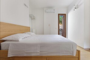 Ein Schlafzimmer mit Holz-Kopfteil, einem Bett mit weißer Bettwäsche, einer Klimaanlage an der Wand und Blick durch eine Tür auf einen Außenbereich.
