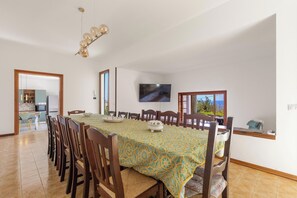 A dining area with a long table, chairs, a television, and a kitchen visible through an open door.