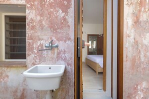 A bathroom with a white sink, a window with a metal grill, and a door leading to a bedroom with a bed and a wooden floor. - Villa Natura by BarbarHouse (Santa Cesarea Terme)