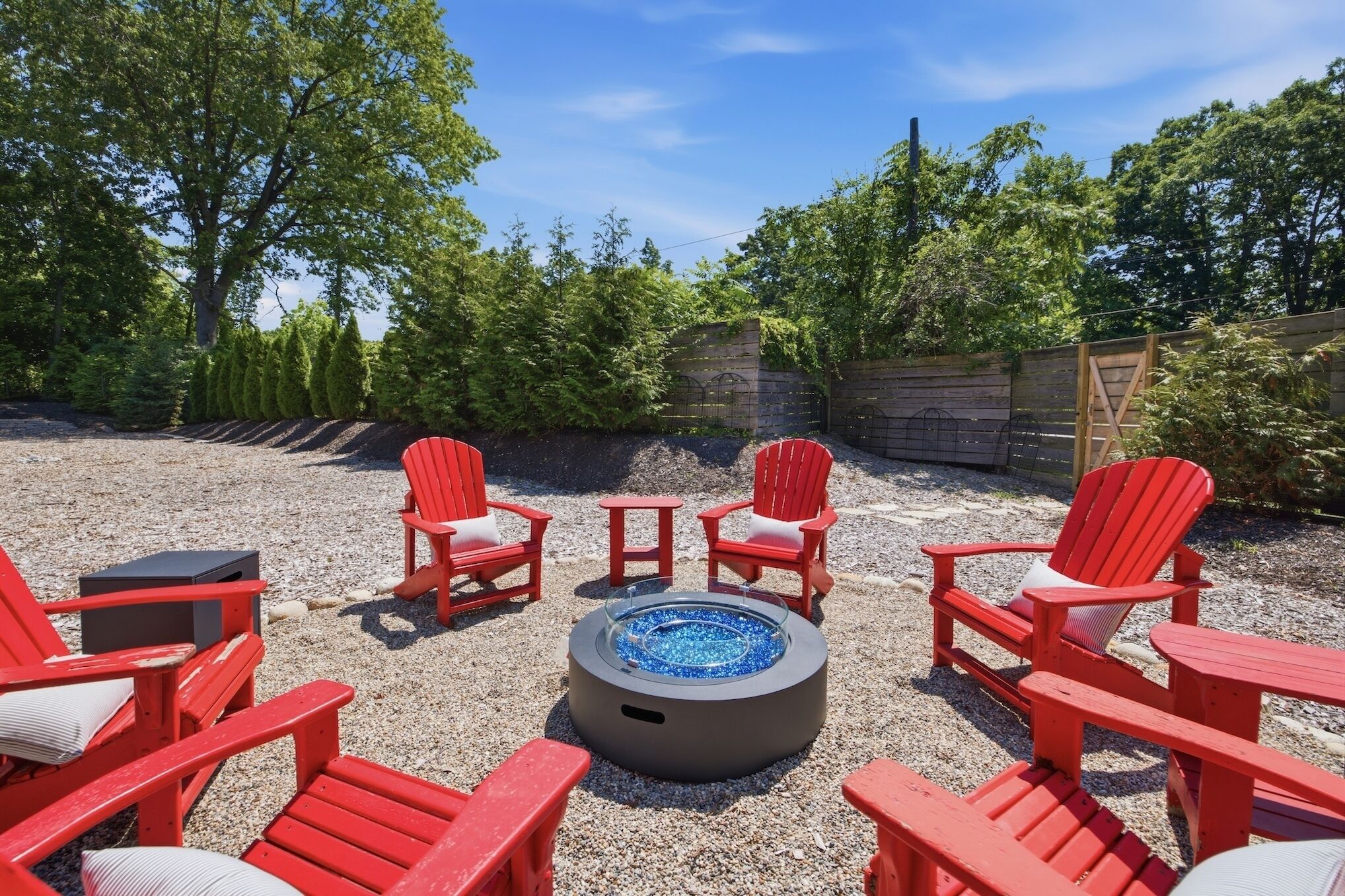 A circular fire pit surrounded by red Adirondack chairs in a gravel area with trees and a wooden fence in the background.