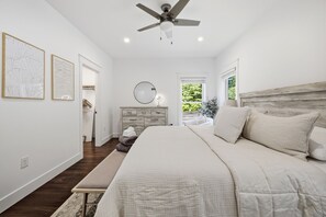A bedroom with a bed, a ceiling fan, a dresser, a mirror, and a window with a view of greenery. - It's A Wonderful Life - Union Pier, MI (New Buffalo)