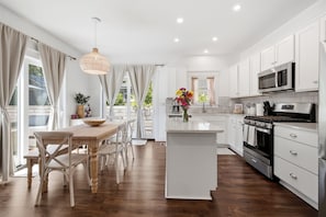 A modern kitchen with white cabinetry, a central island, and a dining area with a wooden table and chairs. - It's A Wonderful Life - Union Pier, MI (New Buffalo)