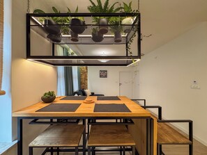A dining area with a wooden table and benches, a hanging light fixture with plants, and a brick wall in the background. - Fully equipped serviced apartment for modern & independent explorers. (Katowice)