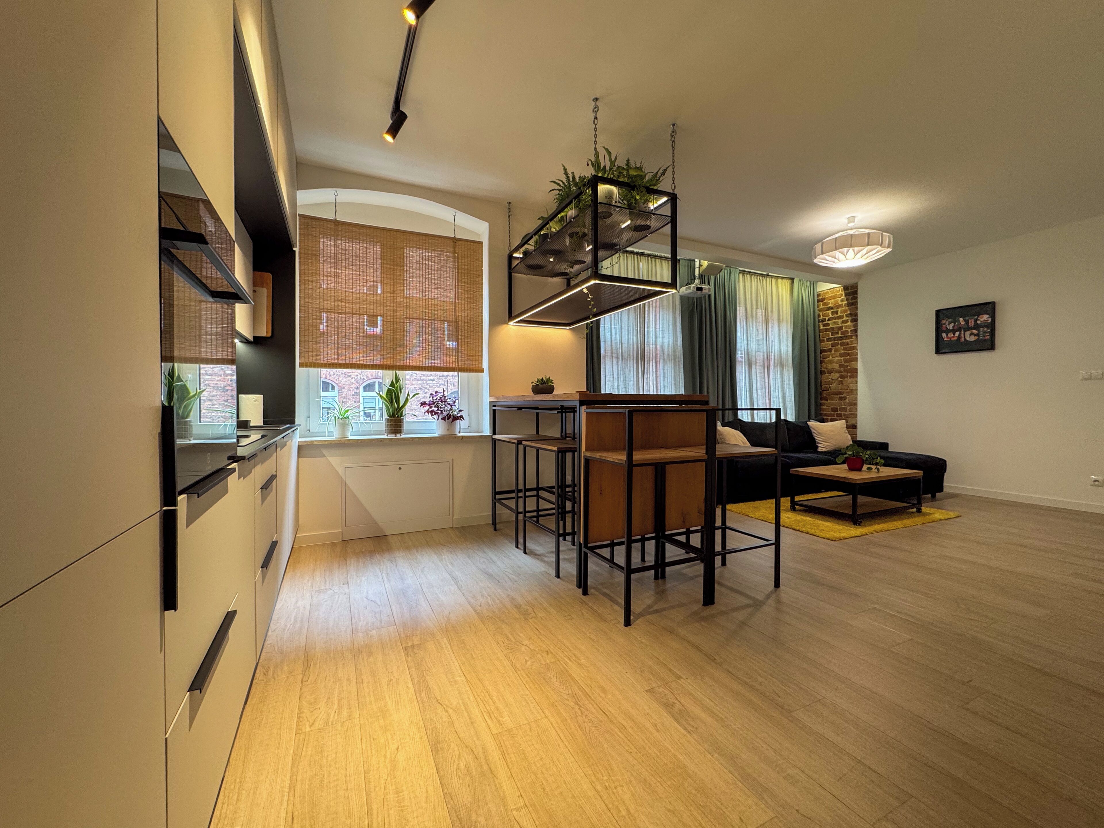 A modern kitchen with a dining area, featuring a bar stool, a sofa, and a hanging plant.