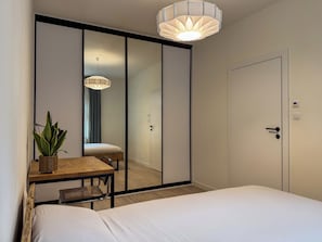 A hotel room with a bed, a desk with a chair, a potted plant, a mirror, and a door with a lock. - Fully equipped serviced apartment for modern & independent explorers. (Katowice)