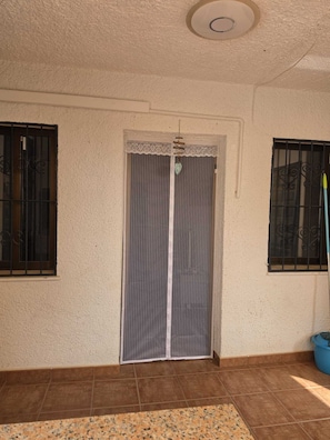 Ein Zimmer mit geschlossener Tür, einem vergitterten Fenster, einem Deckenlüfter und einem blauen Eimer auf dem Boden.